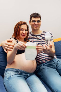 Kanepede otururken kameranın önünde bebek bezi tutan ve besleyen hamile ebeveynlerin gülümsemesi.