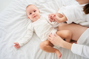 Üzgün, ağlayan küçük bir oğlan çocuğu beyaz büyük bir yatakta yatıyor. Genç bir anne oğlunun bezini değiştirir. Çocuk sağlığı ve bebek bakımı kavramı.