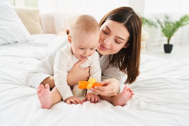 Sevimli genç anne yeni doğmuş tatlı oğluyla yatakta oynuyor, bebeğim. Büyük, parlak bir odada. Annelik ve bakım kavramı.