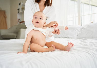 Duştan sonra, yumuşak, kuru bir bezle yatakta oturan yeni doğmuş bir çocuk. Genç bir anne bir bebeği beyaz, parlak bir yatak odasında giydirir. Çocukluk ve annelik kavramı.