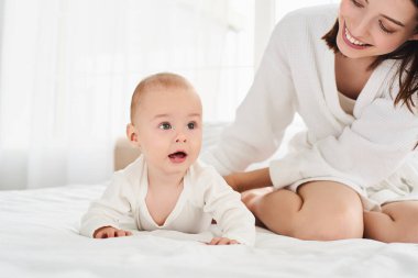 Mutlu, genç, beyaz bir anne, oyun oynuyor, yeni doğmuş oğluna sarılıyor, yeni yürümeye başlamış, bebek, büyük beyaz bir yatakta bornozla, yatak odasında oturuyor..