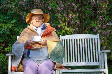 Bahçedeki bankta elinde bir kupa çayla yaşlı bir kadın. Parktaki bankta oturup çay içen mutlu yaşlı kadın. Yaşlılık, bakım, yardım kavramı.