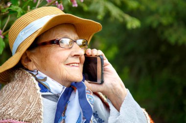 Yaşlı bir kadın parkta bir bankta cep telefonuyla konuşuyor. Yaşlı bir kadın huzurevinin bahçesinde cep telefonu kullanıyor. Teknoloji, yaşlılık ve iletişim kavramı.. 