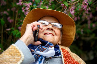Yaşlı bir kadın parkta bir bankta cep telefonuyla konuşuyor. Yaşlı bir kadın huzurevinin bahçesinde cep telefonu kullanıyor. Teknoloji, yaşlılık ve iletişim kavramı.. 
