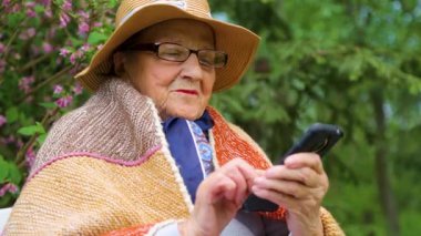 Yaşlı bir kadın parkta bir bankta cep telefonuyla konuşuyor. Yaşlı bir kadın huzurevinin bahçesinde cep telefonu kullanıyor. Teknoloji, yaşlılık ve iletişim kavramı..  