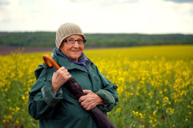 Kolza tohumu tarlasında yaşlı bir kadının portresi. Çiçek açan kolza tohumu tarlasında yürüyen mutlu yaşlı kadın. Dinlenme, rahatlama, mutluluk kavramı.. 