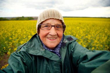 Kolza tohumu tarlasında yaşlı bir kadının portresi. Çiçek açan kolza tohumu tarlasında yürüyen mutlu yaşlı kadın. Dinlenme, rahatlama, mutluluk kavramı.. 