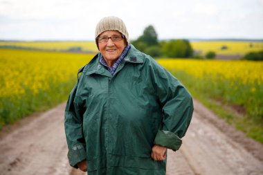 Kolza tohumu tarlasında yaşlı bir kadının portresi. Çiçek açan kolza tohumu tarlasında yürüyen mutlu yaşlı kadın. Dinlenme, rahatlama, mutluluk kavramı.. 