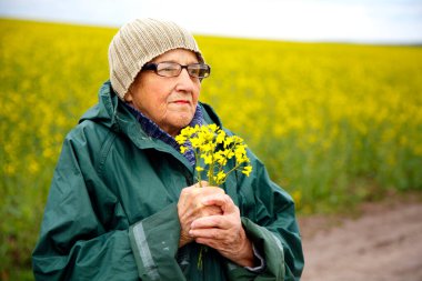Kolza tohumu tarlasında yaşlı bir kadının portresi. Çiçek açan kolza tohumu tarlasında yürüyen mutlu yaşlı kadın. Dinlenme, rahatlama, mutluluk kavramı.. 