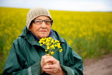 Kolza tohumu tarlasında yaşlı bir kadının portresi. Çiçek açan kolza tohumu tarlasında yürüyen mutlu yaşlı kadın. Dinlenme, rahatlama, mutluluk kavramı.. 