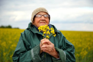 Kolza tohumu tarlasında yaşlı bir kadının portresi. Çiçek açan kolza tohumu tarlasında yürüyen mutlu yaşlı kadın. Dinlenme, rahatlama, mutluluk kavramı.. 