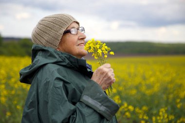Kolza tohumu tarlasında yaşlı bir kadının portresi. Çiçek açan kolza tohumu tarlasında yürüyen mutlu yaşlı kadın. Dinlenme, rahatlama, mutluluk kavramı.. 