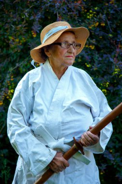 Parkta katanalı bir kimono giymiş yaşlı bir kadın. Bahçede katana ile antrenman yapan yaşlı bir kadının portresi. Yaşlılık, spor, doğa kavramı.. 
