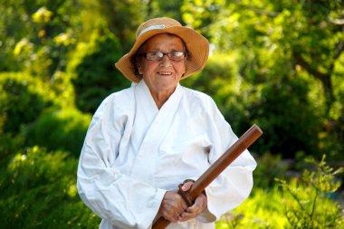 Parkta katanalı bir kimono giymiş yaşlı bir kadın. Bahçede katana ile antrenman yapan yaşlı bir kadının portresi. Yaşlılık, spor, doğa kavramı.. 