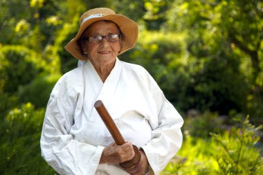 Parkta katanalı bir kimono giymiş yaşlı bir kadın. Bahçede katana ile antrenman yapan yaşlı bir kadının portresi. Yaşlılık, spor, doğa kavramı.. 