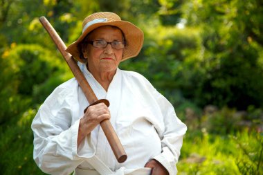 Parkta katanalı bir kimono giymiş yaşlı bir kadın. Bahçede katana ile antrenman yapan yaşlı bir kadının portresi. Yaşlılık, spor, doğa kavramı.. 