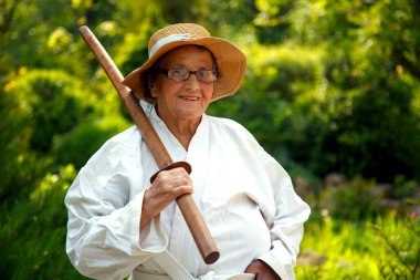 Parkta katanalı bir kimono giymiş yaşlı bir kadın. Bahçede katana ile antrenman yapan yaşlı bir kadının portresi. Yaşlılık, spor, doğa kavramı.. 