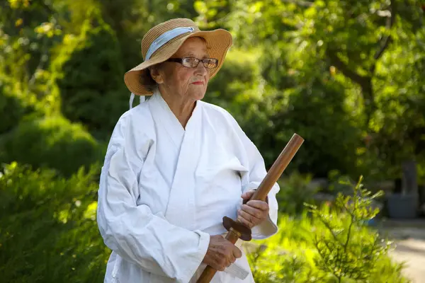 Parkta katanalı bir kimono giymiş yaşlı bir kadın. Bahçede katana ile antrenman yapan yaşlı bir kadının portresi. Yaşlılık, spor, doğa kavramı.. 