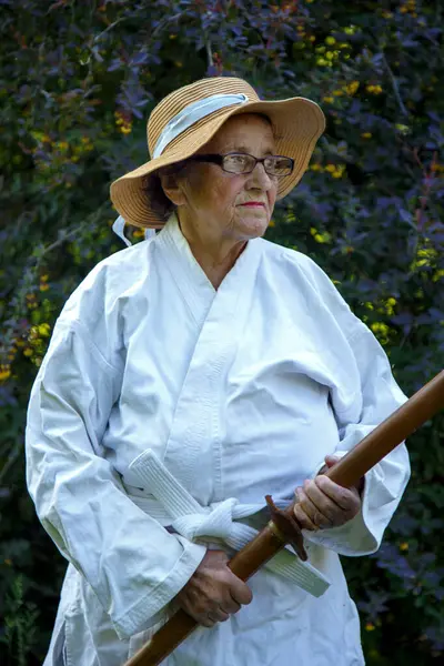 Parkta katanalı bir kimono giymiş yaşlı bir kadın. Bahçede katana ile antrenman yapan yaşlı bir kadının portresi. Yaşlılık, spor, doğa kavramı.. 
