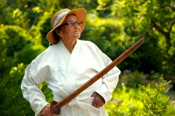 Parkta katanalı bir kimono giymiş yaşlı bir kadın. Bahçede katana ile antrenman yapan yaşlı bir kadının portresi. Yaşlılık, spor, doğa kavramı.. 