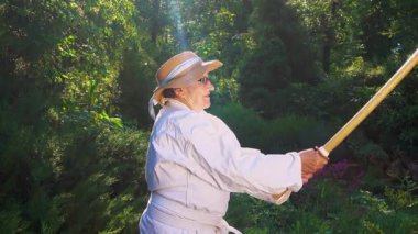Parkta katanalı bir kimono giymiş yaşlı bir kadın. Bahçede katana ile antrenman yapan yaşlı bir kadının portresi. Yaşlılık, spor, doğa kavramı..  
