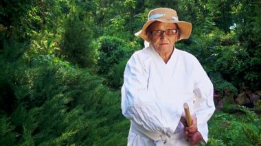 Parkta katanalı bir kimono giymiş yaşlı bir kadın. Bahçede katana ile antrenman yapan yaşlı bir kadının portresi. Yaşlılık, spor, doğa kavramı..  