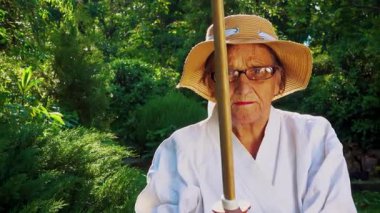 Parkta katanalı bir kimono giymiş yaşlı bir kadın. Bahçede katana ile antrenman yapan yaşlı bir kadının portresi. Yaşlılık, spor, doğa kavramı..  