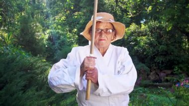 Parkta katanalı bir kimono giymiş yaşlı bir kadın. Bahçede katana ile antrenman yapan yaşlı bir kadının portresi. Yaşlılık, spor, doğa kavramı..  