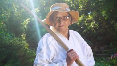 Parkta katanalı bir kimono giymiş yaşlı bir kadın. Bahçede katana ile antrenman yapan yaşlı bir kadının portresi. Yaşlılık, spor, doğa kavramı..  