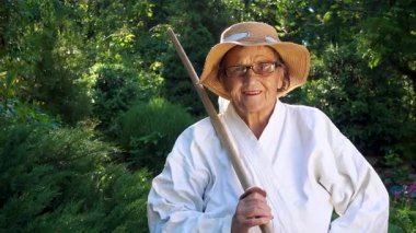 Parkta katanalı bir kimono giymiş yaşlı bir kadın. Bahçede katana ile antrenman yapan yaşlı bir kadının portresi. Yaşlılık, spor, doğa kavramı..  