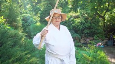 Parkta katanalı bir kimono giymiş yaşlı bir kadın. Bahçede katana ile antrenman yapan yaşlı bir kadının portresi. Yaşlılık, spor, doğa kavramı..  
