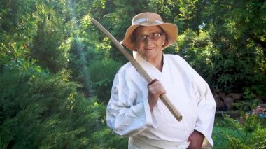 Parkta katanalı bir kimono giymiş yaşlı bir kadın. Bahçede katana ile antrenman yapan yaşlı bir kadının portresi. Yaşlılık, spor, doğa kavramı..  