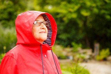 Bahçedeki kameraya poz veren yaşlı güzel bir kadının portresi. Kırmızı pelerinli yaşlı bir kadın pansiyon parkında yürüyor. Yaşlılık, dinlenme, doğa kavramı.. 
