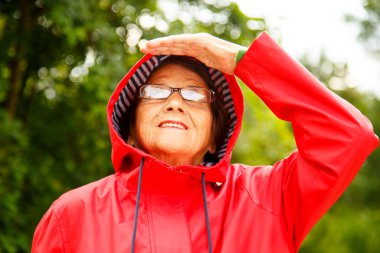 Bahçedeki kameraya poz veren yaşlı güzel bir kadının portresi. Kırmızı pelerinli yaşlı bir kadın pansiyon parkında yürüyor. Yaşlılık, dinlenme, doğa kavramı.. 