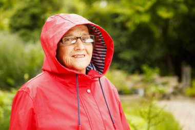 Bahçedeki kameraya poz veren yaşlı güzel bir kadının portresi. Kırmızı pelerinli yaşlı bir kadın pansiyon parkında yürüyor. Yaşlılık, dinlenme, doğa kavramı.. 