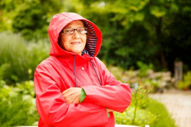 Bahçedeki kameraya poz veren yaşlı güzel bir kadının portresi. Kırmızı pelerinli yaşlı bir kadın pansiyon parkında yürüyor. Yaşlılık, dinlenme, doğa kavramı.. 