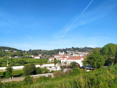 Bereketli yeşil tepelerle çevrili küçük bir kasabanın manzarası açık mavi gökyüzünün altında dağınık bulutlarla, binaların ve doğanın bir karışımını gösteriyor..