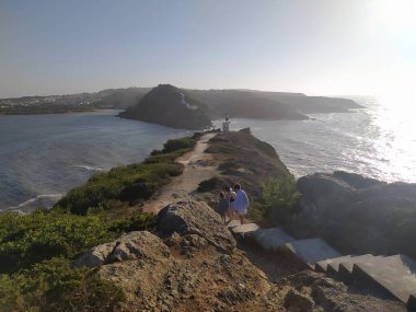 Deniz feneri kıyı şeridi yakınlarındaki kayalık araziye tünemiş, güneş batarken ve okyanus dalgaları üzerinde altın bir parıltı yayarken, sakin bir atmosfer yaratıyor..