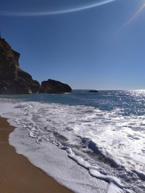 Güneş sakin bir sahilin üzerinde ışıl ışıl parlıyor. Yumuşak dalgalar kumlu sahile dokunuyor, dramatik bir uçurum tarafından çerçeveleniyor, sakin bir sahil atmosferi yaratıyor..