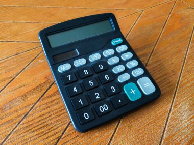 Hafif tozlu, siyah bir hesap makinesi sıcak kahverengi ahşap bir yüzeye dayanır. Basit tasarımı ve net görüntüsü onu finans, matematik veya günlük hesaplamalar kavramlarını resmetmek için mükemmel kılar..