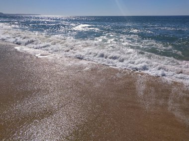 Kumlu sahil, yumuşak dalgaların parlak ve berrak bir gökyüzünün altında kıyıya ulaştığı sahil boyunca uzanır ve ziyaretçiler için huzurlu bir atmosfer yaratır..
