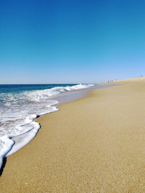 Kumlu sahil, yumuşak dalgaların parlak ve berrak bir gökyüzünün altında kıyıya ulaştığı sahil boyunca uzanır ve ziyaretçiler için huzurlu bir atmosfer yaratır..