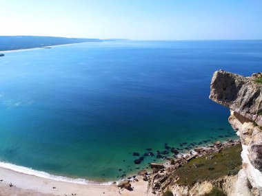 Geniş kıyı manzarası berrak mavi sular ve kayalık bir uçurumla çevrili huzurlu bir kumsal olduğunu gösteriyor. Güneş dingin manzarayı aydınlatıyor, sakin bir atmosfer yaratıyor..