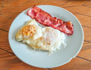 Kızarmış yumurta ve sade beyaz bir tabakta mükemmel pişirilmiş pastırma. Klasik bir kahvaltı yemeği resmi, menüler, bloglar ya da mutfak siteleri için mükemmel. Yüksek kalite, iştah açıcı fotoğrafçılık.