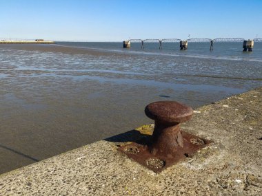 Dalgaların çekildiği sakin bir yere bakan beton bir iskelede kırsal, yıpranmış bir boğa. Uzak yapılar derinlik ekler. Seyahat, kıyı veya deniz teması için ideal.