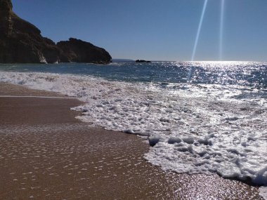 Güneş sakin bir sahilin üzerinde ışıl ışıl parlıyor. Yumuşak dalgalar kumlu sahile dokunuyor, dramatik bir uçurum tarafından çerçeveleniyor, sakin bir sahil atmosferi yaratıyor..