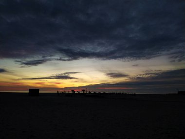 Sıcak, bulutlu bir gökyüzüne karşı plaj yapılarını siluetleyen dramatik sahil batımı. Bitişlerin, başlangıçların, umudun ve sükunetin temaları için mükemmel..