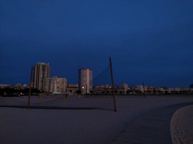 Sahilde sakin bir alacakaranlık sahnesi, şehrin ufuk çizgisine karşı boş bir voleybol ağı..