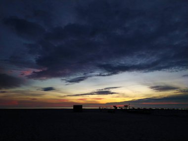 Üzerinde kara bulutlar olan dramatik plaj günbatımı, ufku boyayan canlı renkler. Siluetli yapılar derinlik gizemi ekler. Seyahat, doğa ya da atmosferik ruh hali için mükemmel..