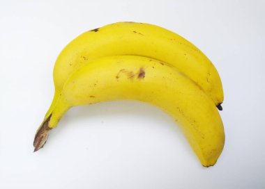 İki olgun muz, ince benekli, temiz beyaz arkaplana karşı nazik bir eğri oluşturur. Basit ama çarpıcı bir görüntü, sağlıklı beslenme, tropikal temalar veya minimalist tasarım projeleri için mükemmel..